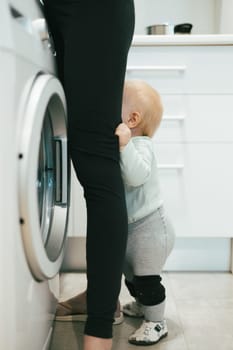 Little infant baby boy child hiding between mothers legs demanding her attention while she is multitasking, trying to do some household chores in kitchen at home. Mother on maternity leave