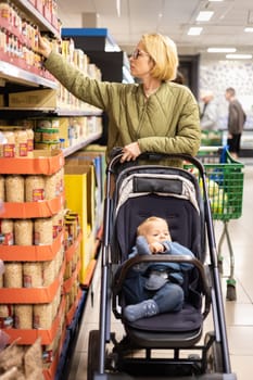 Casualy dressed mother choosing canned products in department of supermarket grocery store with her infant baby boy child in stroller