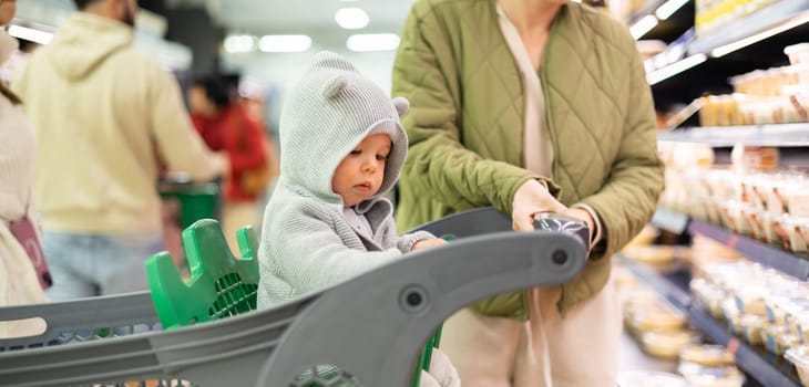Casualy dressed mother choosing products in department of supermarket grocery store with her infant baby boy child in shopping cart