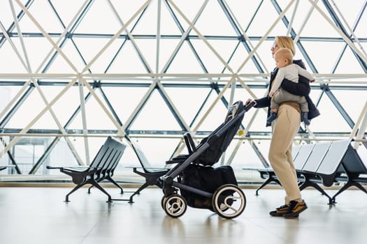 Mother carying his infant baby boy child, pushing stroller at airport departure terminal moving to boarding gates to board an airplane. Family travel with baby concept