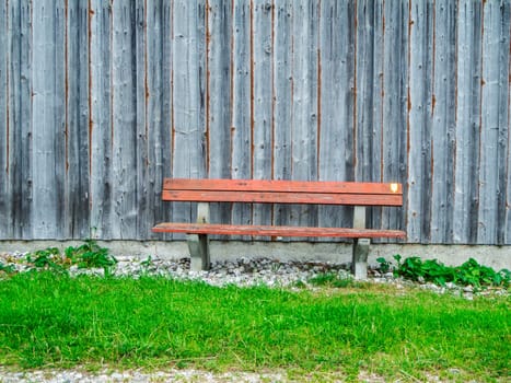 Frontal view of a barn wall with vertical boards and a weathered park bench in front of it and a small lawn in Bavaria.