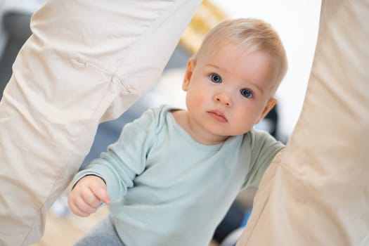 Portrait of adorable curious infant baby boy child taking first steps holding to father's pants at home. Cute baby boy learning to walk