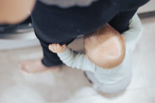 Top down view of little infant baby boy hiding between mothers legs demanding her attention while she is multitasking, trying to do some household chores in kitchen at home. Mother on maternity leave