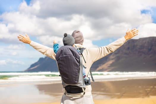 Young father rising hands to the sky while enjoying pure nature carrying his infant baby boy son in backpack on windy sandy beach of Famara, Lanzarote island, Spain. Family travel concept