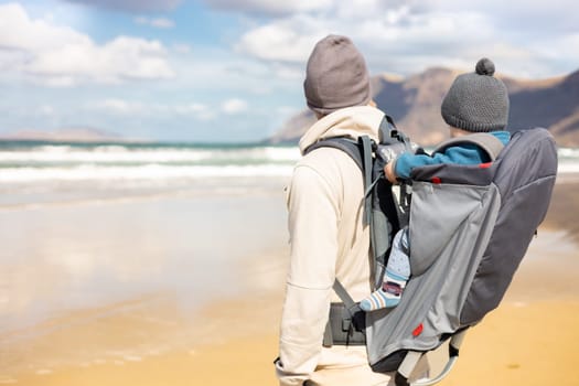 Young father carrying his infant baby boy son in backpack on windy sandy beach of Famara, Lanzarote island, Spain. Family travel and winter vacation concept. Parenting, fatherhood, father's day
