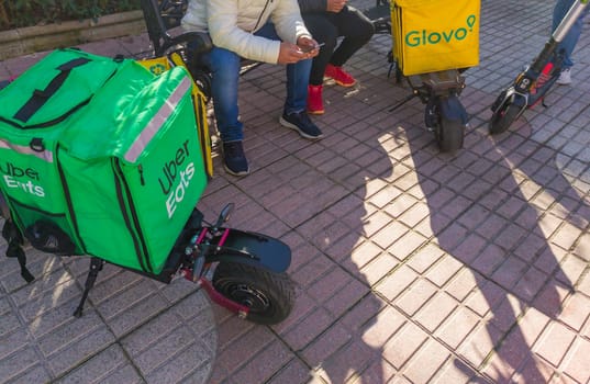 Caceres, Spain - Jan 19th, 2021: Delivery service backpacks and electric kick scooters. Urban background
