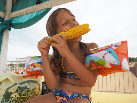 Anapa, Russia, August 19, 2021. Caucasian girl 7 years old eats hot boiled corn on the beach on a summer day. A hungry child after bathing with wet hair holds a cob. Sun canopy on a sandy beach.