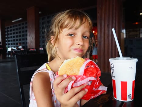 Anapa Russia 23 August 2021 A beautiful girl of 7 years old is eating at the KFC restaurant, enjoying the food and smiling. Caucasian child holding a burger. Cola in a cardboard cup with lid and straw