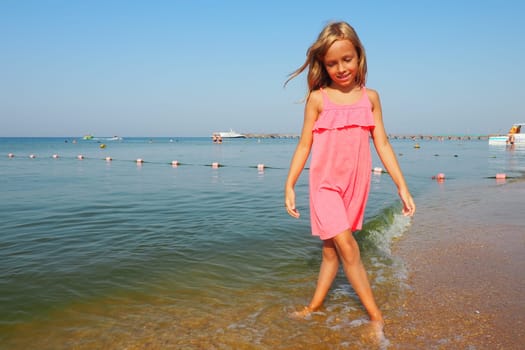Anapa, Russia, August 29, 2021 A 7-year-old girl in a pink dress walks along the sea. Blond hair and sunburn on the skin. The girl smiles and looks down. Sea wave, swimming people, boats and blue sky.