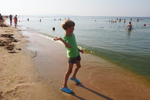 Anapa, Russia, August 29, 2021. A 9-year-old boy throws a stone, shell or coin into the sea. Caucasian child with blond hair in green clothes. Sun, sky, sea and swimming tourists. Throwing an object