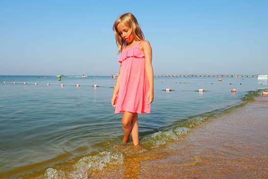 Anapa, Russia, August 29, 2021. A 7-year-old girl in a pink dress walks along the sea. Blond hair and sunburn on the skin. The girl looks down. Sea waves, buoys, swimming people, boats and blue sky.