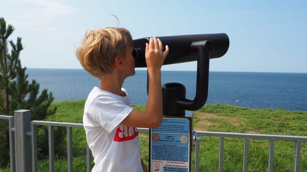 Anapa, Russia, August 23, 2021 A boy looks through an outdoor telescope. A 9-year-old Caucasian child holds a large marine telescope with his hands. The street of the resort town and walking tourists.