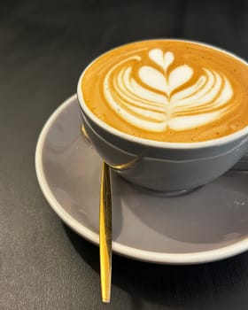 Latte art coffee in a grey ceramic cup with a golden spoon on a saucer, resting on a dark surface