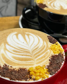 Two cups of cappuccino decorated with latte art, chocolate flakes and biscuit crumbs, served on a wooden table in a cafe