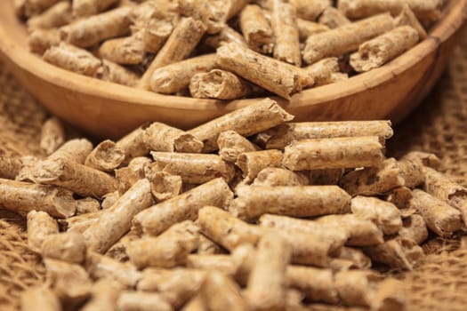 Close-up of a pile of wood pellets in front of and inside a wooden bowl on jute fabric. Focus on the wood pellets in the foreground with warm, natural colours.