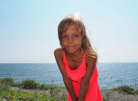 A young pretty girl of 8 years old with blond long hair, tanned skin smiles against the backdrop of a sea landscape. Pink sundress dress. Sunbeams counterlight on a child's hair. Blue sky and horizon