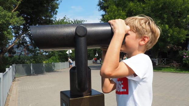 Anapa, Russia, August 23, 2021 A boy looks through an outdoor telescope. A 9-year-old Caucasian child holds a large marine telescope with his hands. The street of the resort town and walking tourists.