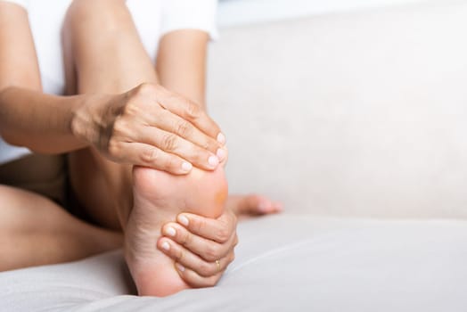 Foot pain, Asian woman sitting on sofa feeling pain in her foot at home, female suffering from feet ache use hand massage relax muscle from soles in home interior, Healthcare and podiatry medical