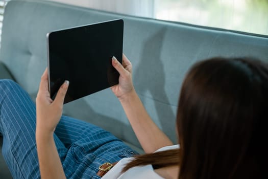 Happy Asian woman playing smart tablet at home while relaxing on sofa living room, Beautiful female sitting on couch using digital tablet pc, technology social media