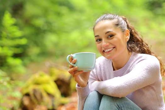 Happy woman sitting in nature holding coffee cup posing looking at camera