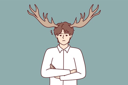 Disgruntled guy with deer horns on head stands with arms crossed and looks at screen with displeasure. Metaphor of man who suffered from infidelity of girlfriend or adultery wife