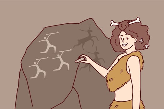 Ancient woman in clothing made from animal skins and bones in hair makes rock paintings. Girl depicts people hunting deer on giant stone and smiling looks at screen