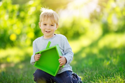 Smiling child sitting on the spring meadow with model of the green house in his hands. Concept of the real estate, rental and buying of a new apartment.