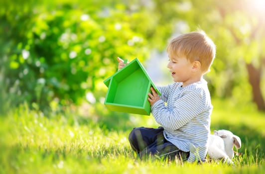 Smiling child sitting on the spring meadow with model of the green house in his hands. Concept of the real estate, rental and buying of a new apartment.