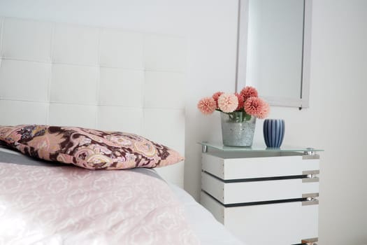 Bedroom interior design. Mirror on the wall. A bouquet of beautiful pink flowers on a white table in the bedroom. Interior Design. Bed with white upholstered headboard. Decorative pillow.