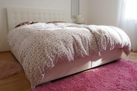 Cozy compact bedroom with white furniture pink carpet and tulle curtains. Fluffy feather blanket is thrown over the bed. Romantic morning. Double bed with upholstered headboard. Light from the window