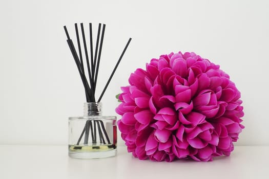 Arrangement with incense sticks, essential oil in a glass vase and a bright pink-lilac terry artificial flower in the interior of a white room. White background.