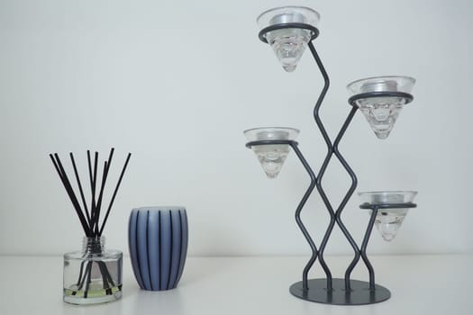 Tall metal chandelier with glass holders for small candles. White background. Incense sticks in a glass cubic vessel with essential oil. Interior decor. Scented gray striped candle or vase