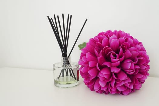 Arrangement with incense sticks, essential oil in a glass vase and a bright pink-lilac terry artificial flower in the interior of a white room. White background.