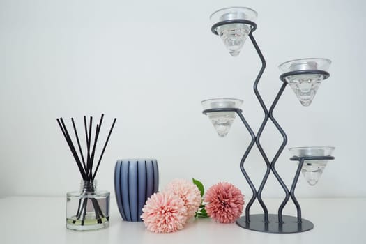 Composition with incense sticks, essential oil in a glass vase and pink terry artificial flowers. Tall metal chandelier with glass holders for small candles. Scented gray striped candle or vase.