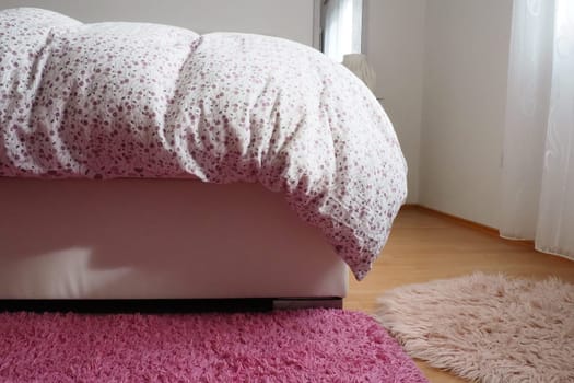 Cozy compact bedroom, white furniture, pink carpet and tulle curtains. Fluffy feather blanket thrown over the double bed. Romantic morning. Light from the window. The blanket hangs down to the floor