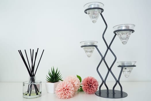 Composition with incense sticks, essential oil in a glass vase and pink terry artificial flowers. Tall metal chandelier. Scented gray striped candle or vase. Artificial green plant aloe or kalanchoe.
