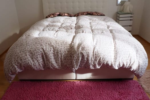 Cozy compact bedroom with white furniture pink carpet and tulle curtains. Fluffy feather blanket is thrown over the bed. Romantic morning. Double bed with upholstered headboard. Light from the window