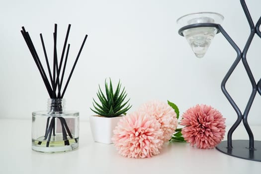 Composition with incense sticks, essential oil in a glass vase and pink terry artificial flowers. Tall metal chandelier. Scented gray striped candle or vase. Artificial green plant aloe or kalanchoe.