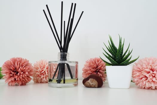 Composition of pink artificial flowers, incense sticks in a glass vase with essential oil. Still life on a white table. Aloe flower in a white pot. Room and apartment decor, interior design