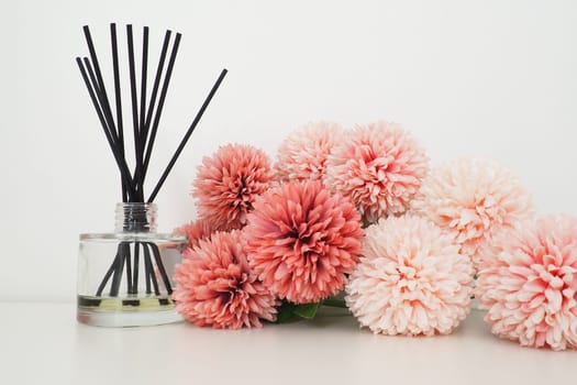 Composition with incense sticks, essential oil in a glass vase and pink terry artificial flowers in the interior of a white room. White background. Lots of flowers and scent sticks