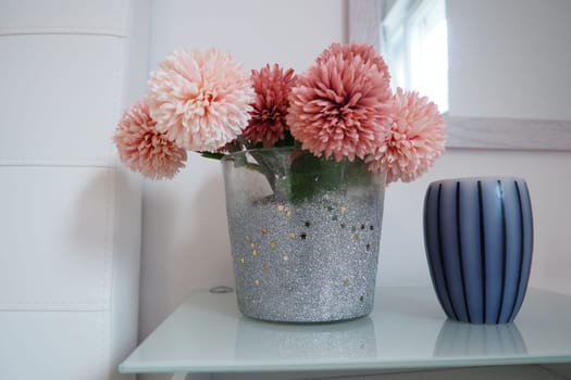 A bouquet of beautiful pink artificial flowers on a white table in the bedroom. Interior Design. Interior design of the women bedroom. Bouquet in a vase. Aromatic striped candle. Mirror on the wall
