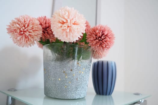 A bouquet of beautiful pink artificial flowers on a white table in the bedroom. Interior Design. Interior design of the women bedroom. Bouquet in a vase. Aromatic striped candle. Mirror on the wall