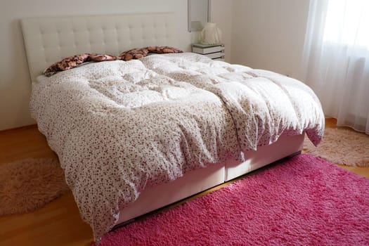 Cozy compact bedroom with white furniture, pink carpet and tulle curtains. Fluffy feather blanket is thrown over the bed. Romantic morning. Double bed with upholstered headboard