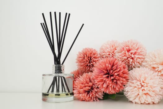 Composition with incense sticks, essential oil in a glass vase and pink terry artificial flowers in the interior of a white room. White background. Lots of flowers and scent sticks