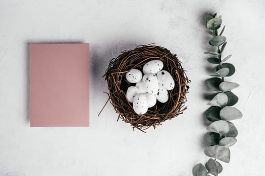 Easter composition of pink blank empty postcard on white gray paper background and white small spot quail eggs in bird brown natural nest near branch of green eucalyptus. Celebrate traditional holiday