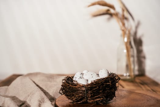 Easter composition of quail spot eggs in nest on wooden table and branch of wheat in glass on beige white blurred background. Celebration of traditional event feast holiday. Rustic comfort style