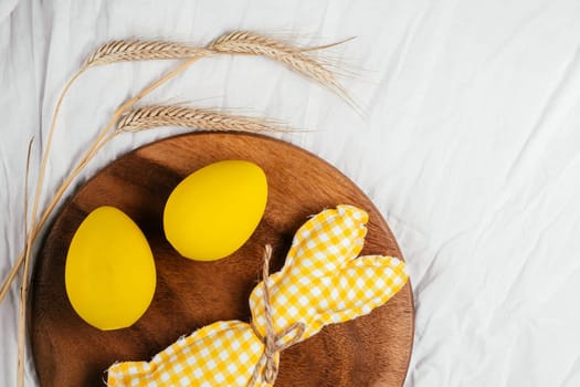 Easter postcard for celebration holiday, one handmade square brown, white and yellow colored bunny with string-scarf on white gray textile background, yellow chicken eggs in wooden plate near cereals