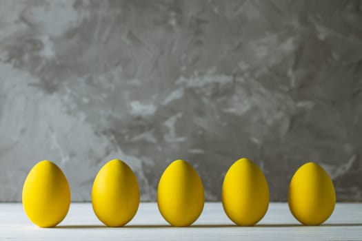 Row of five bright yellow Easter eggs, standing on white wooden surface on gray concrete background, copy space
