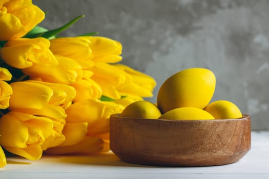 Easter eggs in wooden bowl and bouquet of bright yellow tulips on white wooden surface on gray background, copy space
