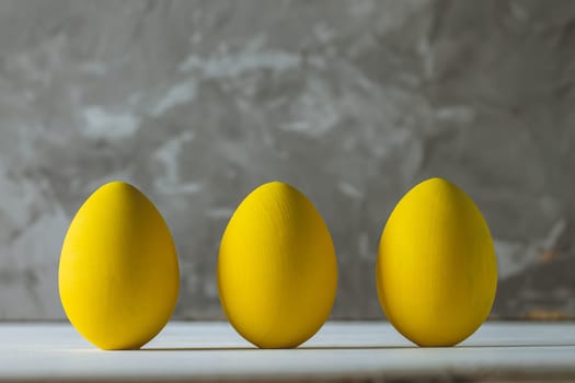 Row of five bright yellow Easter eggs, standing on white wooden surface on gray concrete background, copy space